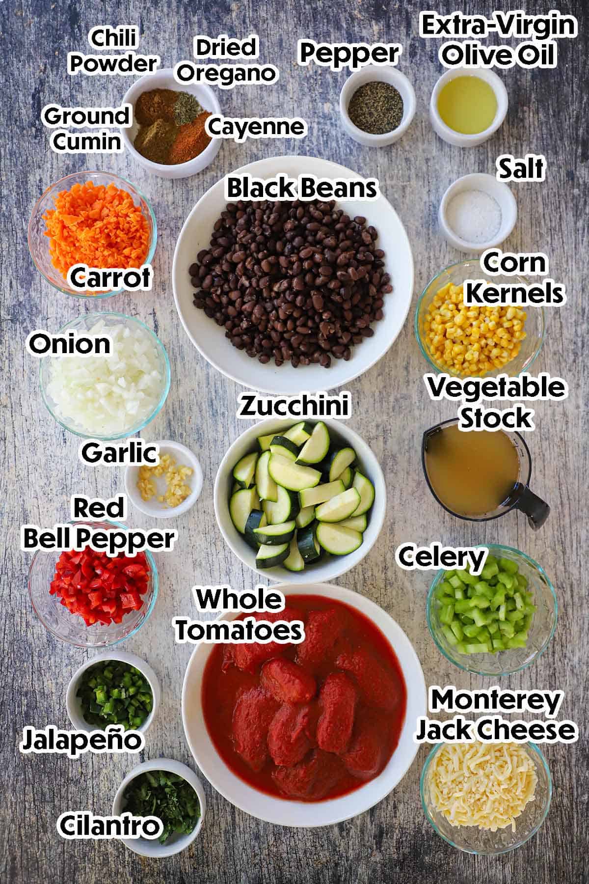 An arrangement of ingredients for Vegetable Chili with Black Beans on a grey wooden background including zucchini, corn, black beans, carrots, onions, tomatoes, peppers, celery, cheese, oil, and seasonings.