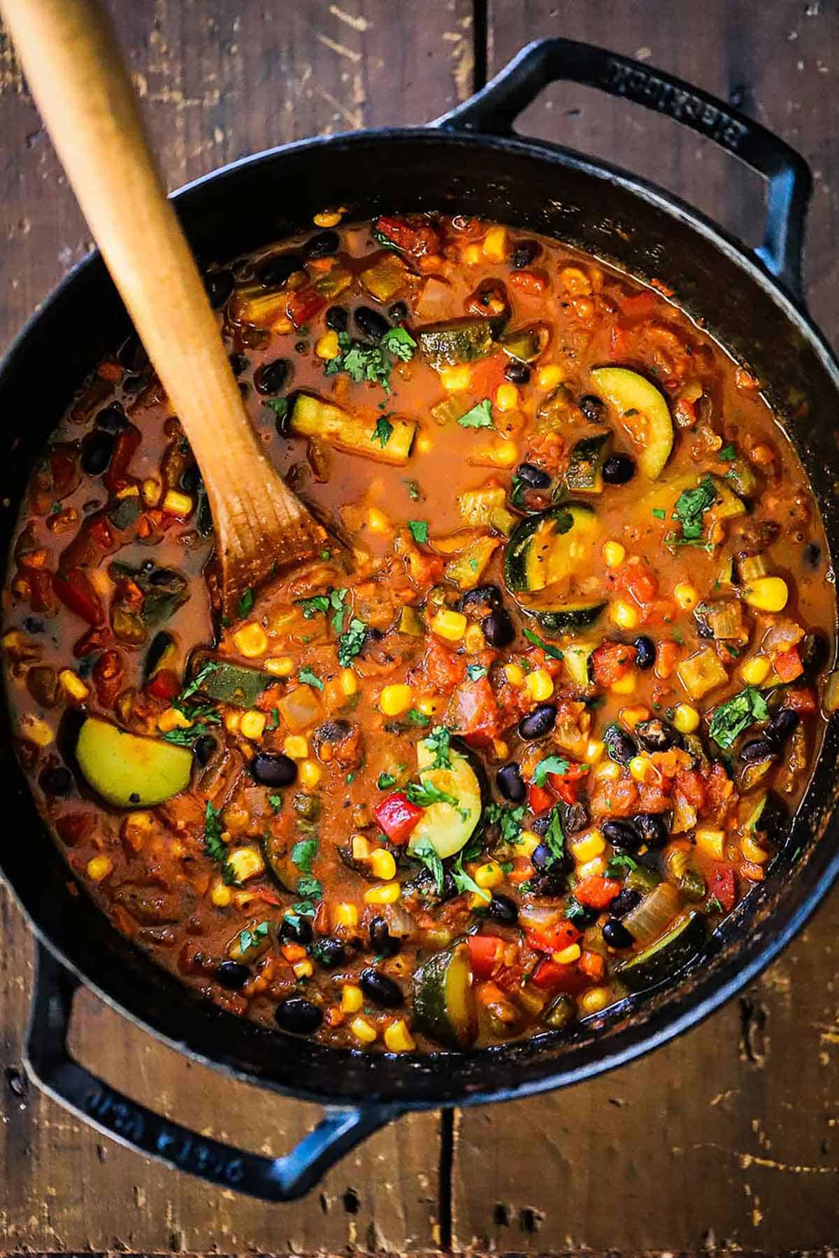 An overhead view of a large black cast-iron pot filled with simmering Vegetable Chili with Black Beans on a gas stove.