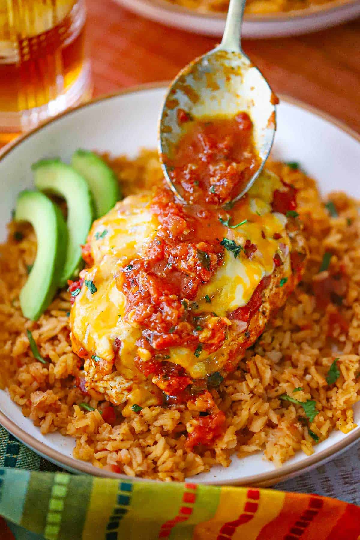 A person using a spoon to drizzle a baked salsa sauce over the top of Easy Baked Salsa Chicken resting on a bed of Mexican rice in a shallow bowl.