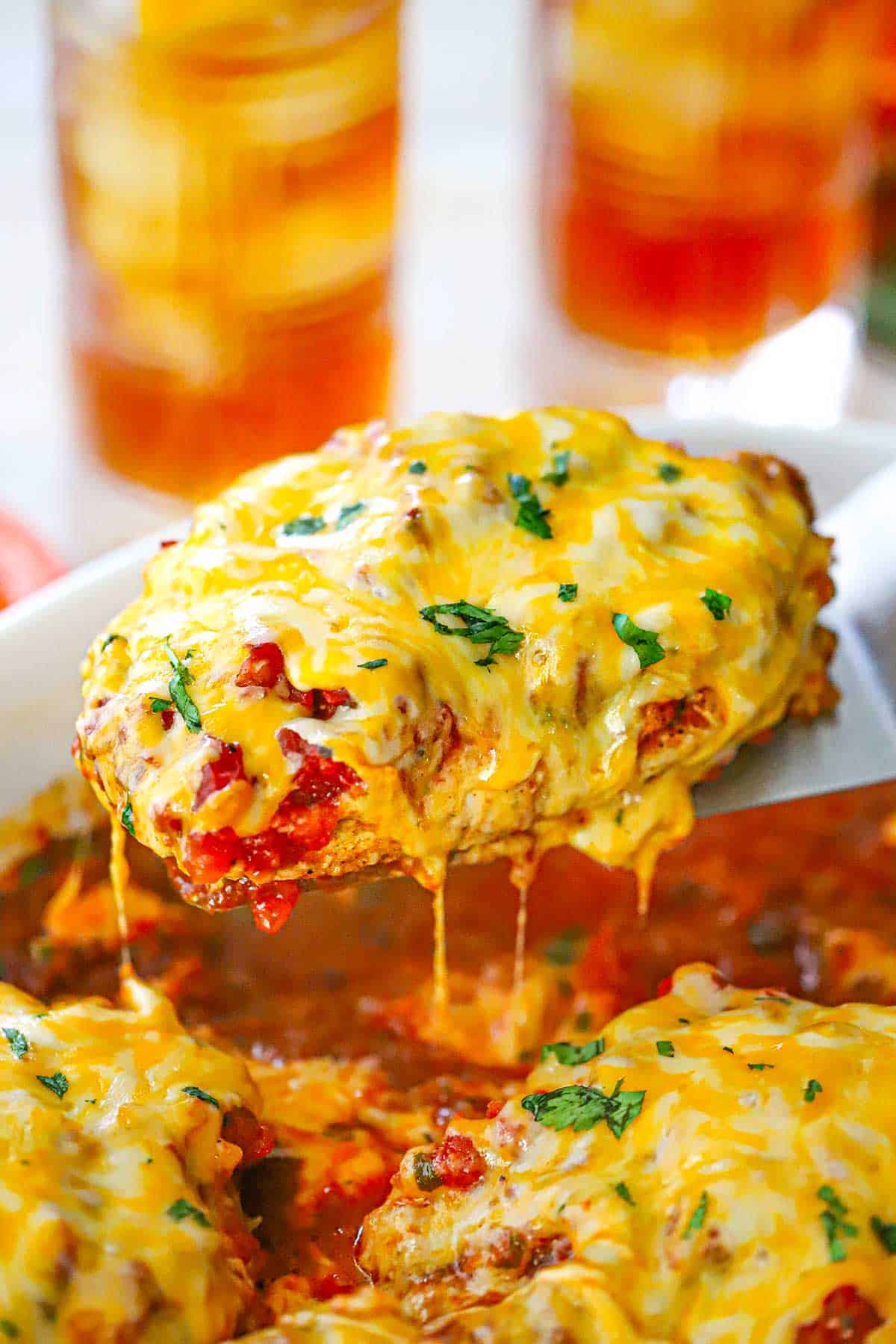 A person using a metal spatula to lift a cheesy salsa chicken breast from a casserole dish holding more of the same.