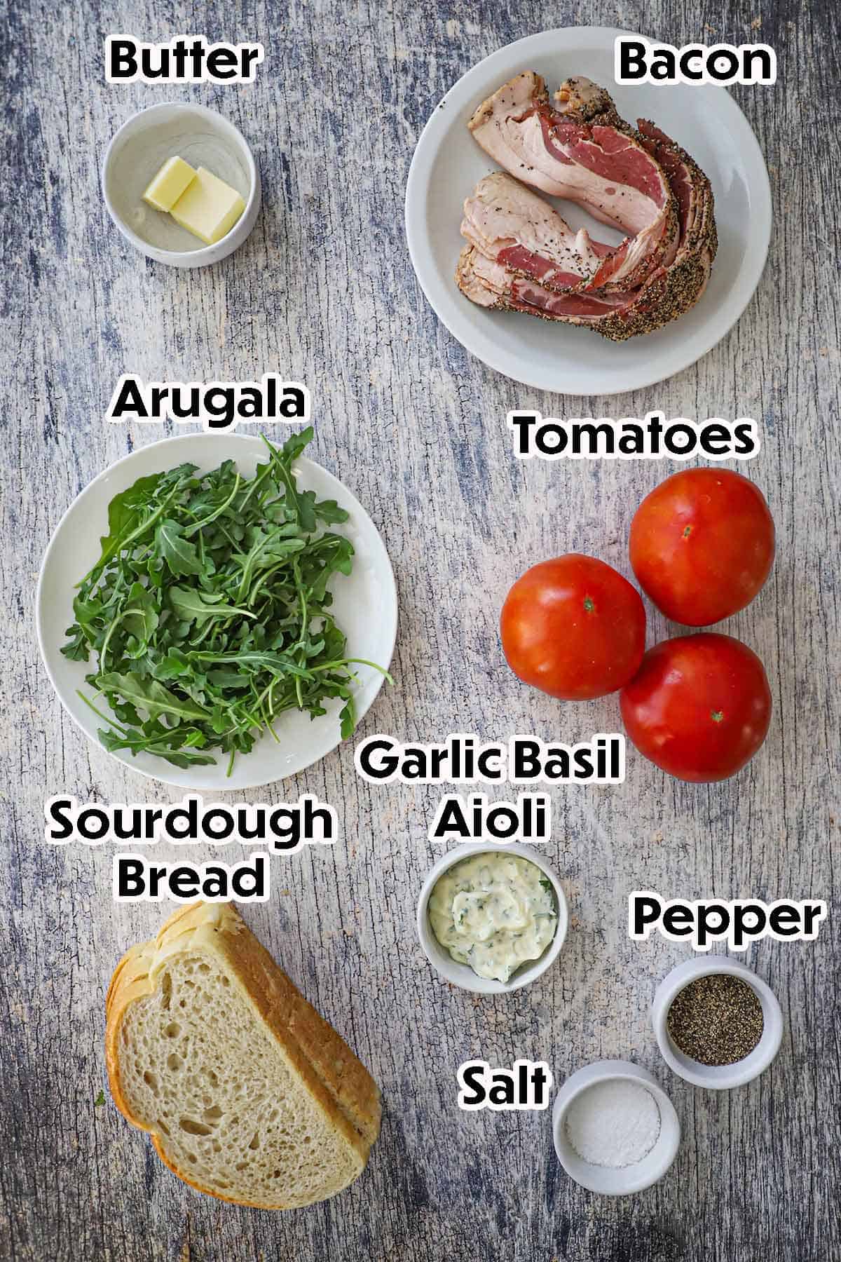 An arrangement of ingredients for Classic BLT with Garlic Basil Aioli on a grey wooden background including peppered bacon, tomatoes, arugula, sourdough bread, butter, garlic basil aioli, salt, and pepper.