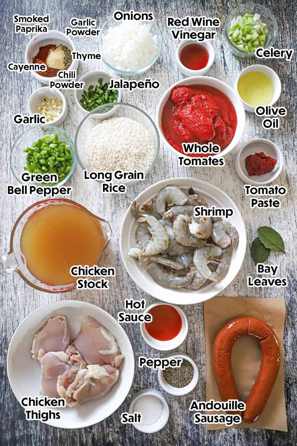 An arrangement of ingredients for authentic jambalaya on a grey wooden background including shrimp, chicken, sausage, rice, tomatoes, peppers, onions, garlic, celery, tomato paste, chicken stock, and seasonings.