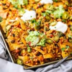 A close-up view of Sheet Pan Nachos topped with melty cheese, vegetarian chili, sliced pickled jalapeños, and dollops of sour cream and homemade guacamole