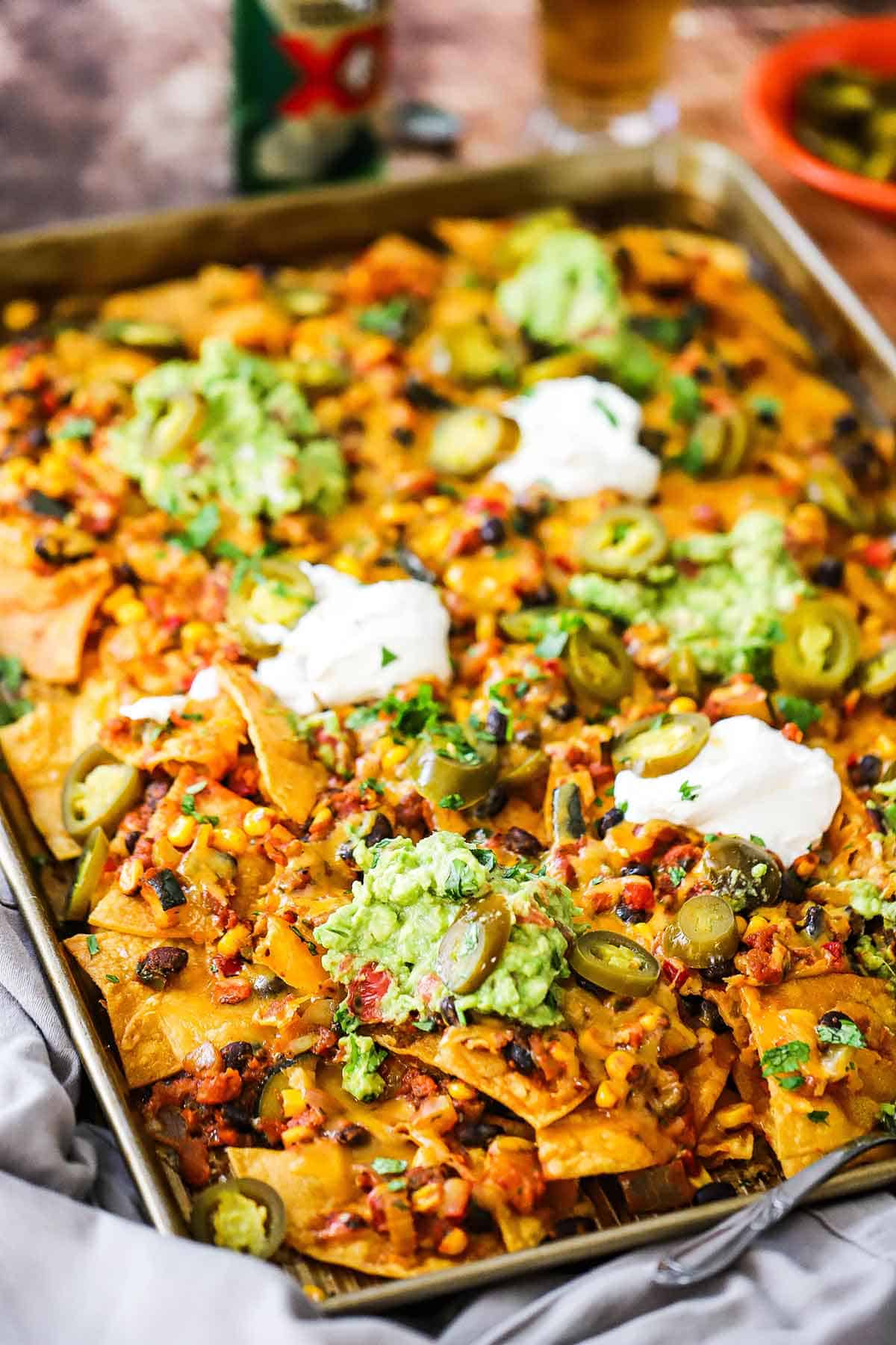 A straight-on view of Sheet Pan Nachos topped with melty cheese, vegetarian chili, sliced pickled jalapeños, and dollops of sour cream and homemade guacamole.