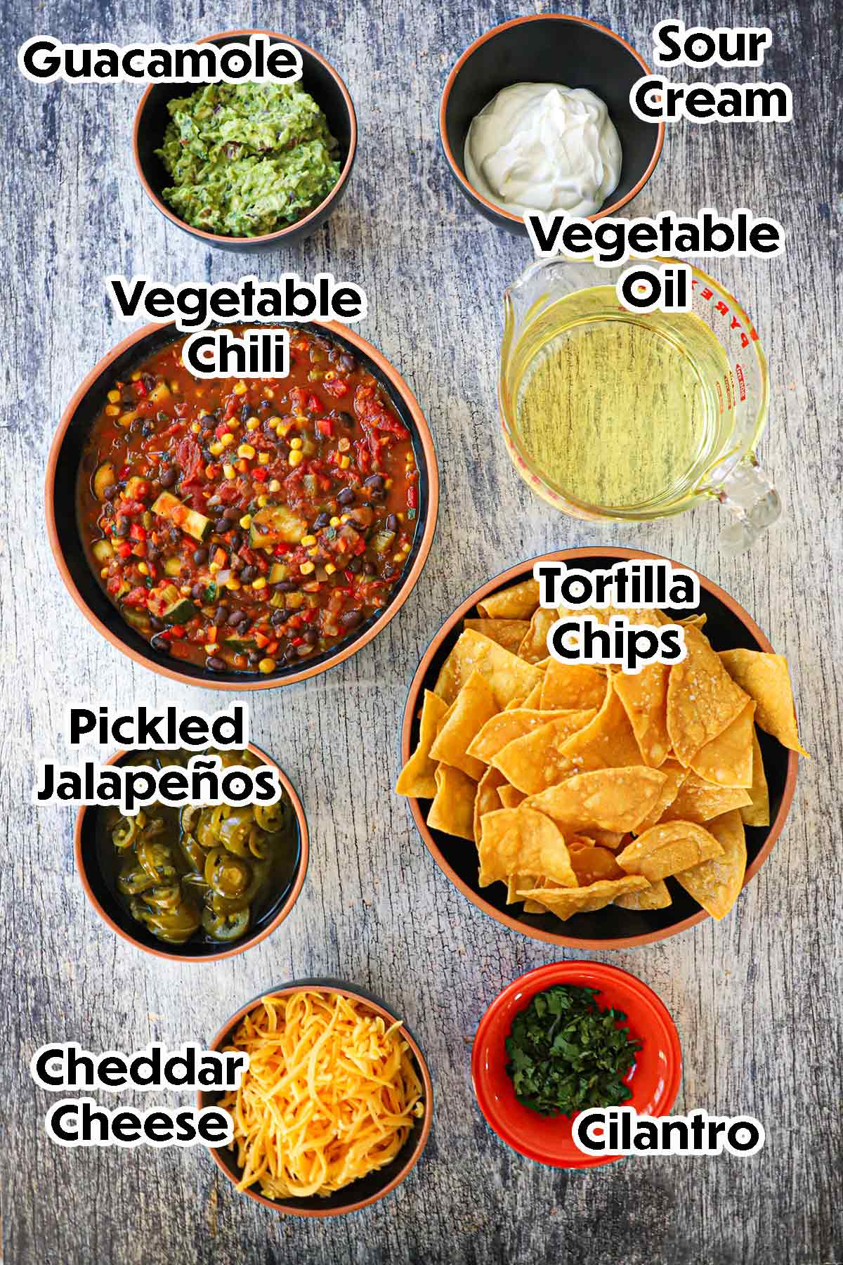 An arrangement of ingredients for Sheet Pan Nachos on a grey wooden background including tortilla chips, vegetable chili, cheddar cheese, oil, guacamole, sour cream, jalapeños, and cilantro.