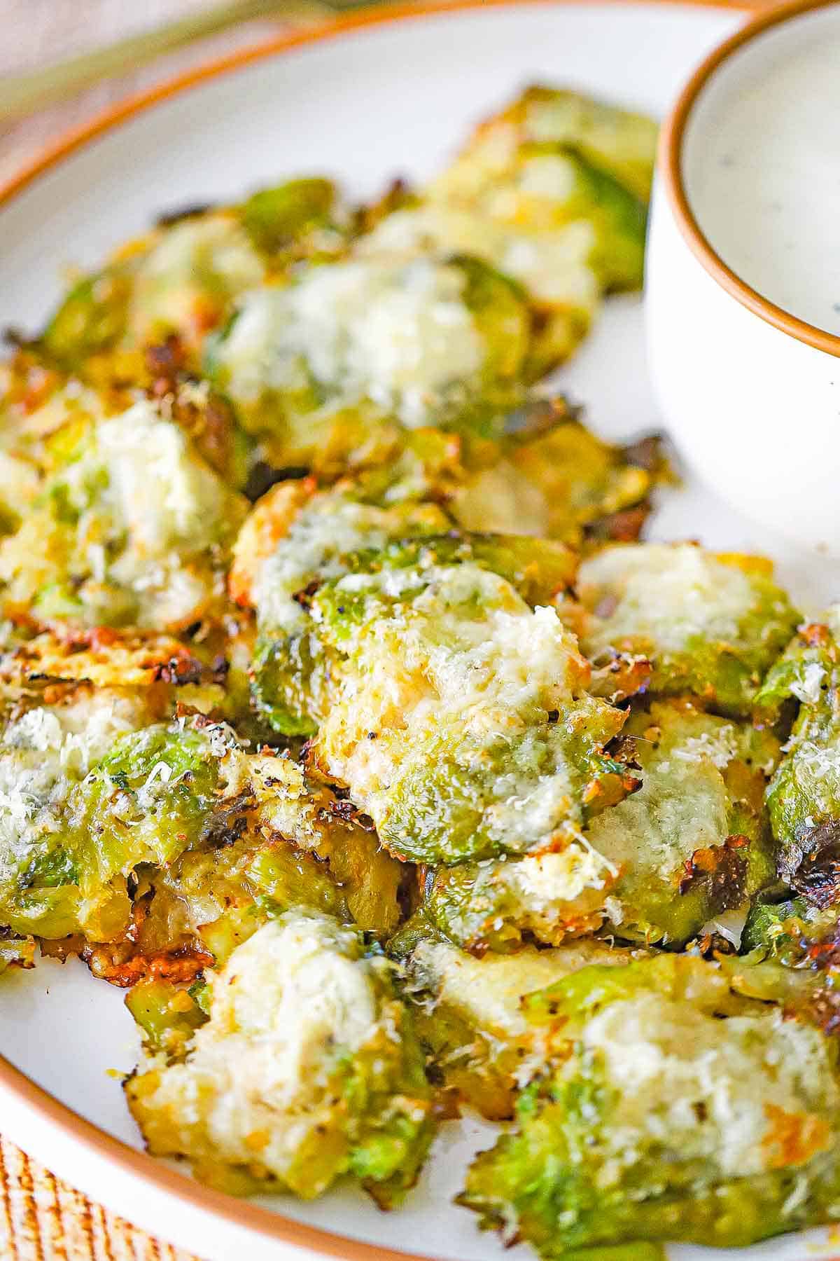 A close-up view of a pile of Parmesan Smashed Brussels Sprouts on a white plate with a small bowl of Ranch dressing nearby.