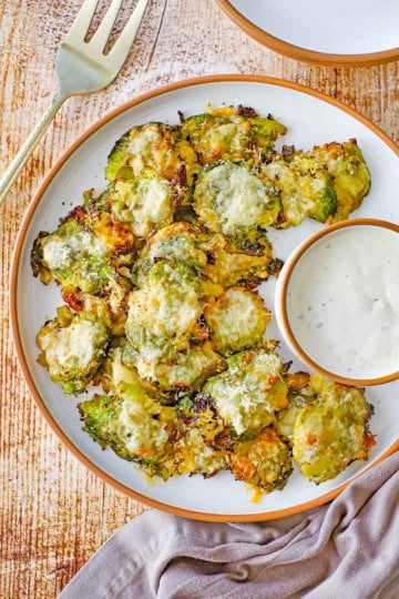 An overhead view of a round white plate filled with a serving of Parmesan Smashed Brussels Sprouts in a pile next to a round bowl filled with homemade Ranch dressing.
