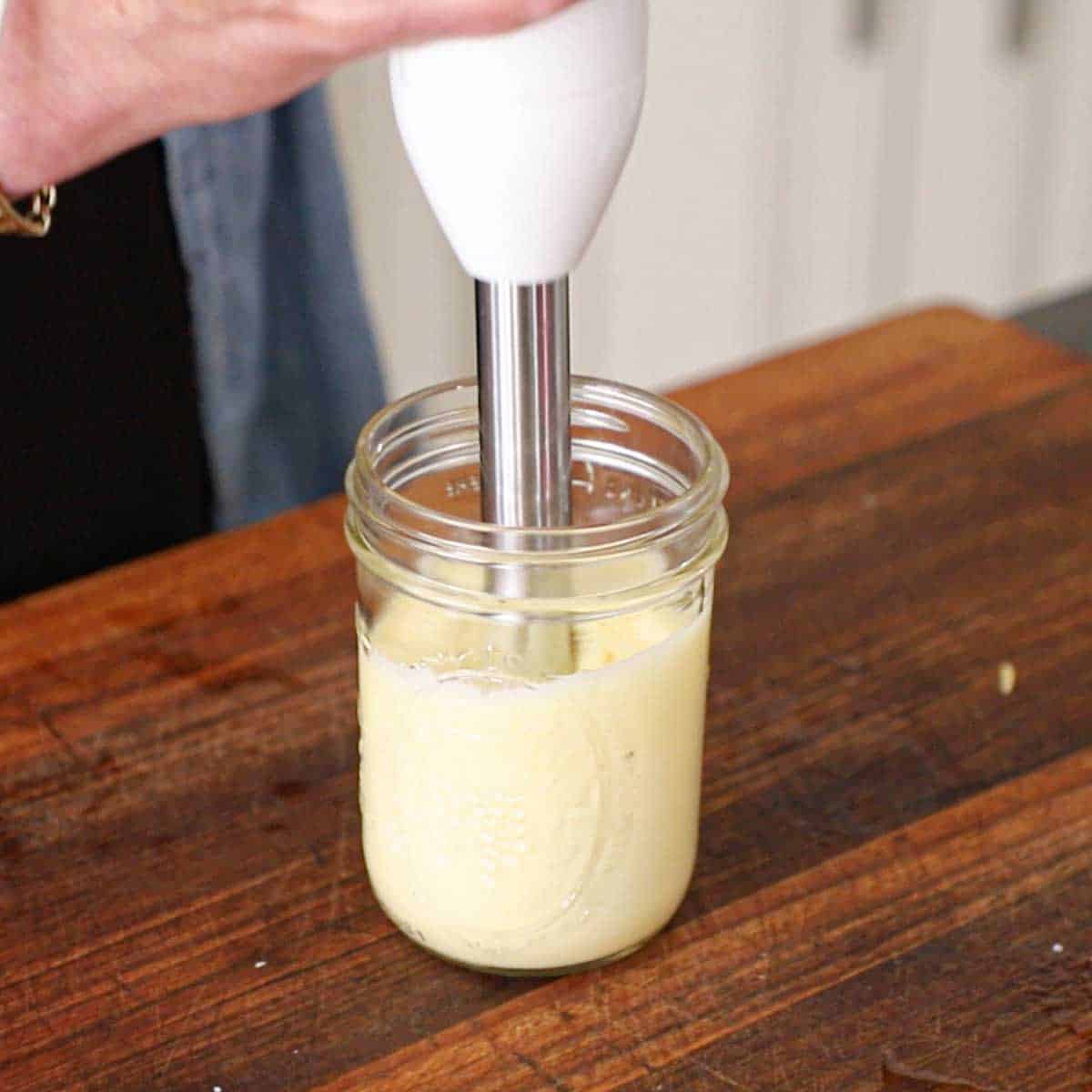 A person using an immersion blender to create homemade mayonnaise in a glass jar on a wooden cutting board.