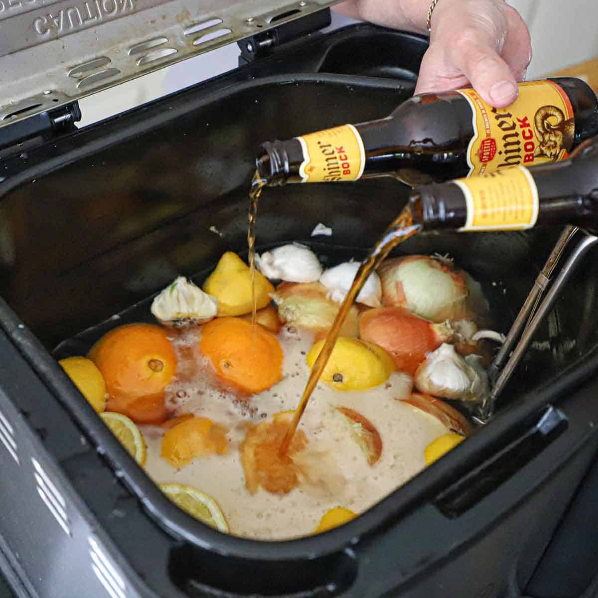 A person pouring beer from two bottles into a large turkey fryer that is filled with water, fruit, onions, and garlic.