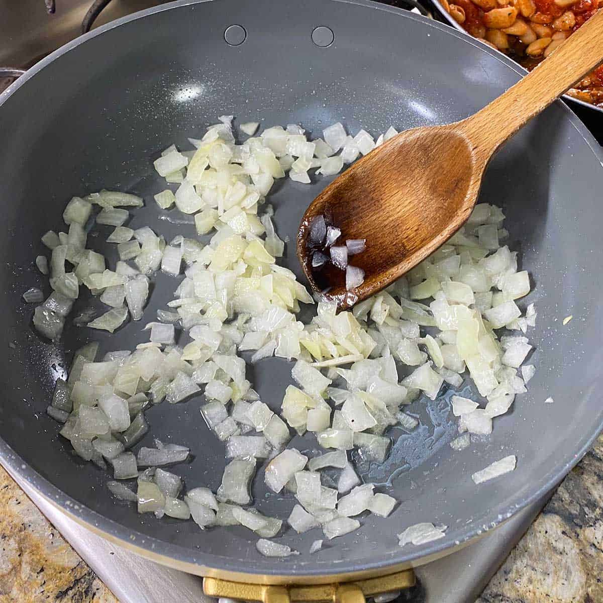 A person using a wooden spoon to stir and sauté chopped onions in olive oil in a large non-stick skillet on a gas stove.