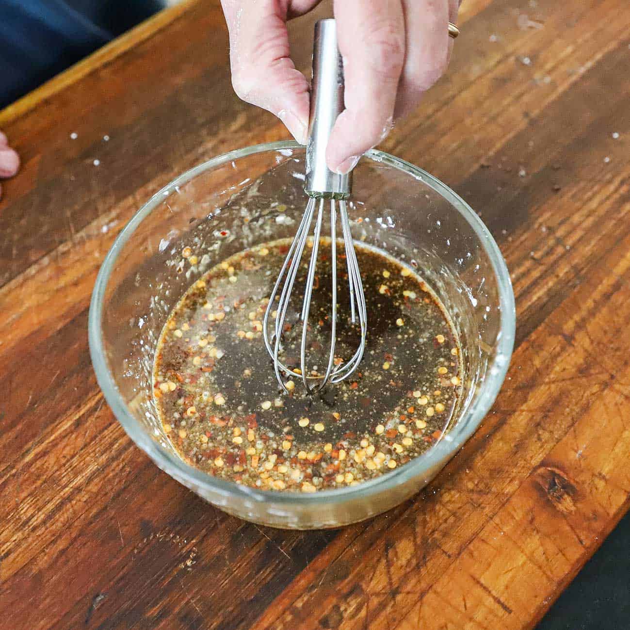 A person using a small whisk to mix soy sauce with brown sugar, vinegar, sesame oil, red pepper flakes, and cornstarch in a glass bowl on a wooden cutting board.