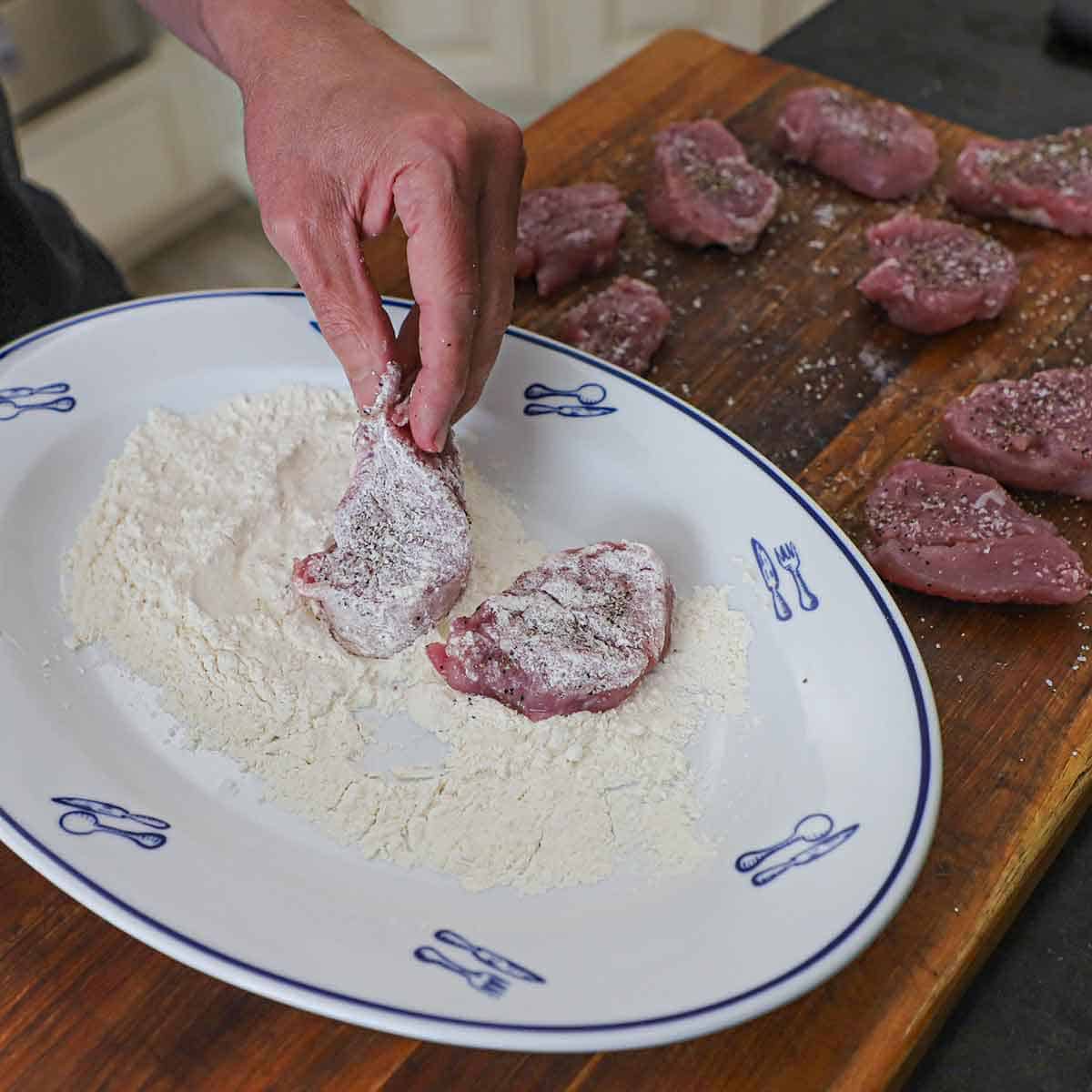 A person using his hands to dredge seasoned pork medallions in a flour in a large oval platter on a cutting board.