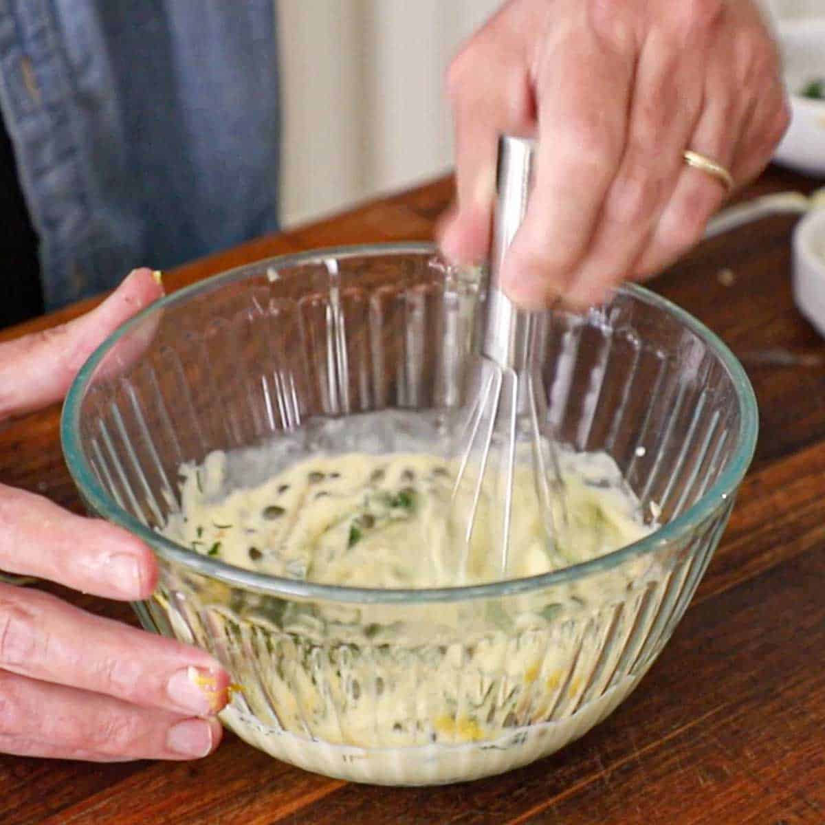 A person using a whisk to combine all the components of Lemon-Dill and Caper Aioli in a glass bowl on a wooden cutting board.