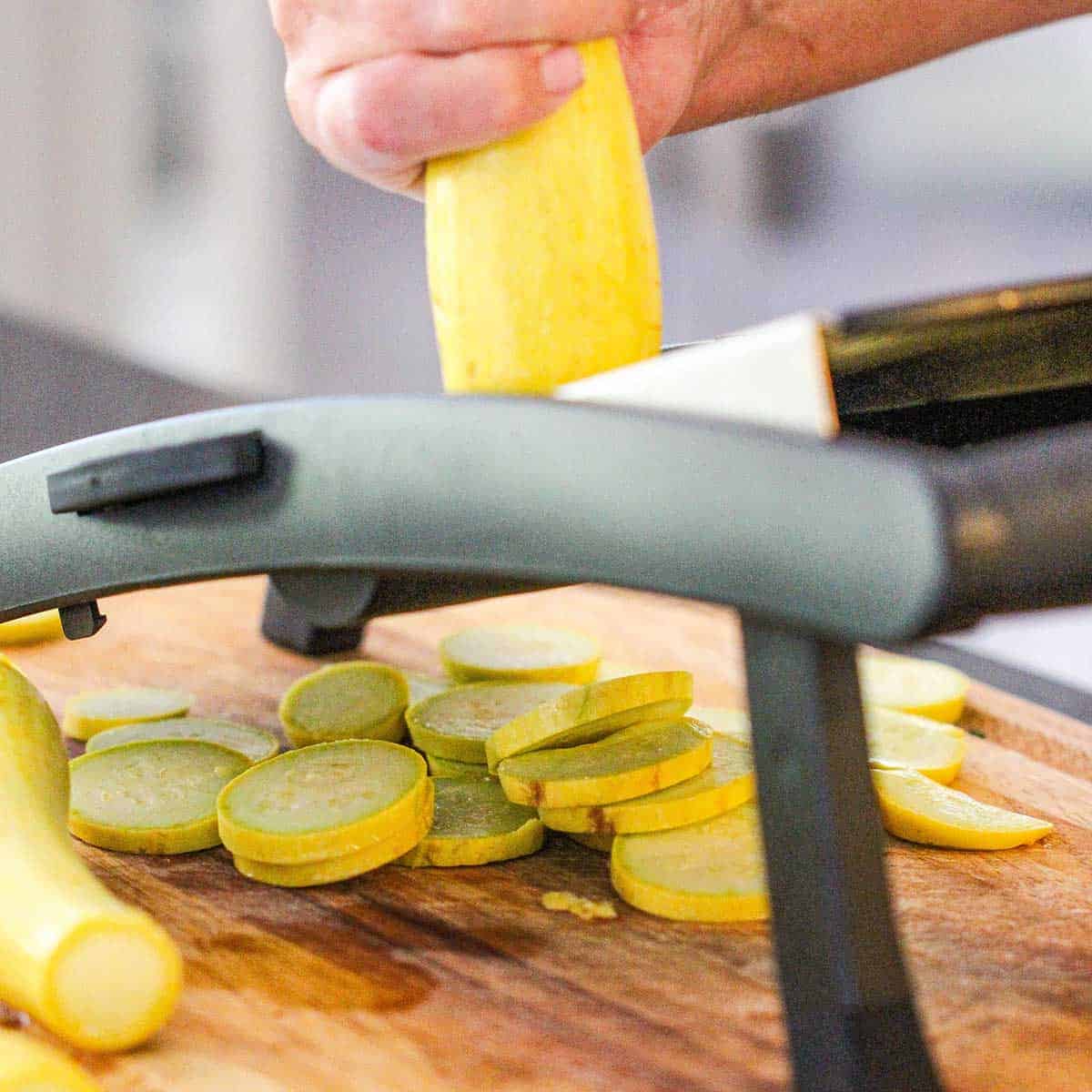 A person using a mandolin to slice a yellow squash into quarter-inch rounds onto a wooden cutting board.