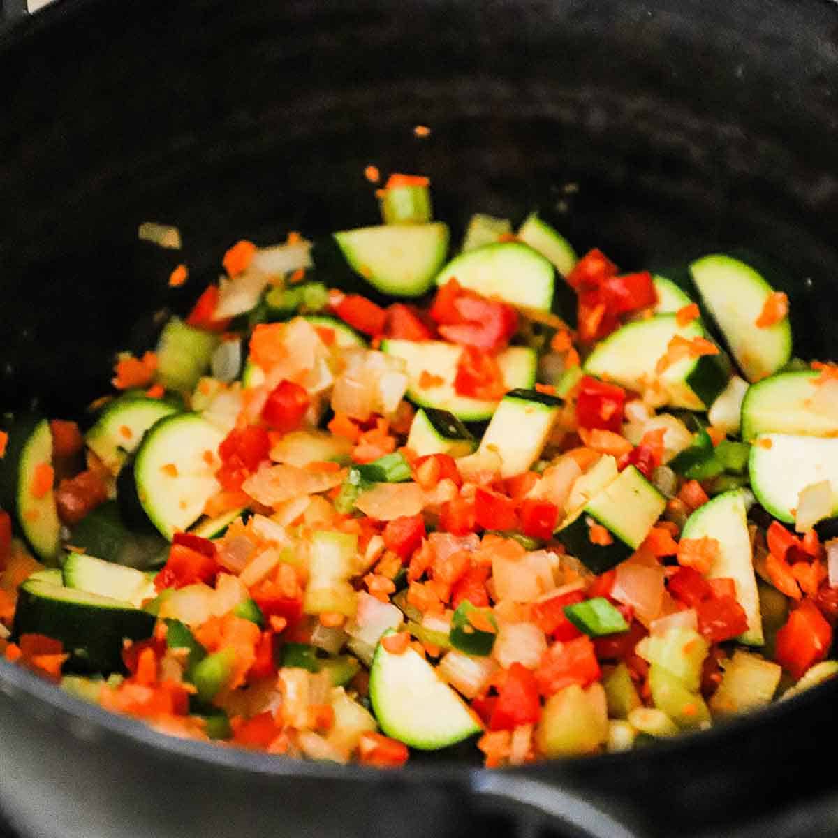 A close-up view of chopped onion, bell peppers, zucchini, celery, carrot, jalapeño, and garlic being sautéed in olive oil in a cast-iron skillet.