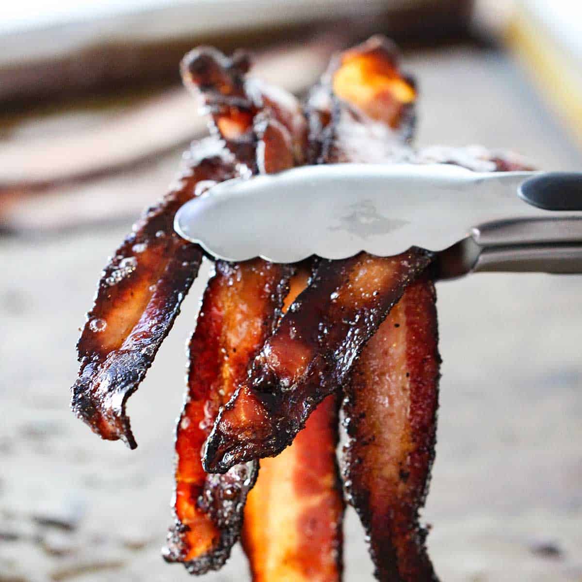 A person using a pair of metal tongs to hold up several strips of crispy black pepper bacon.