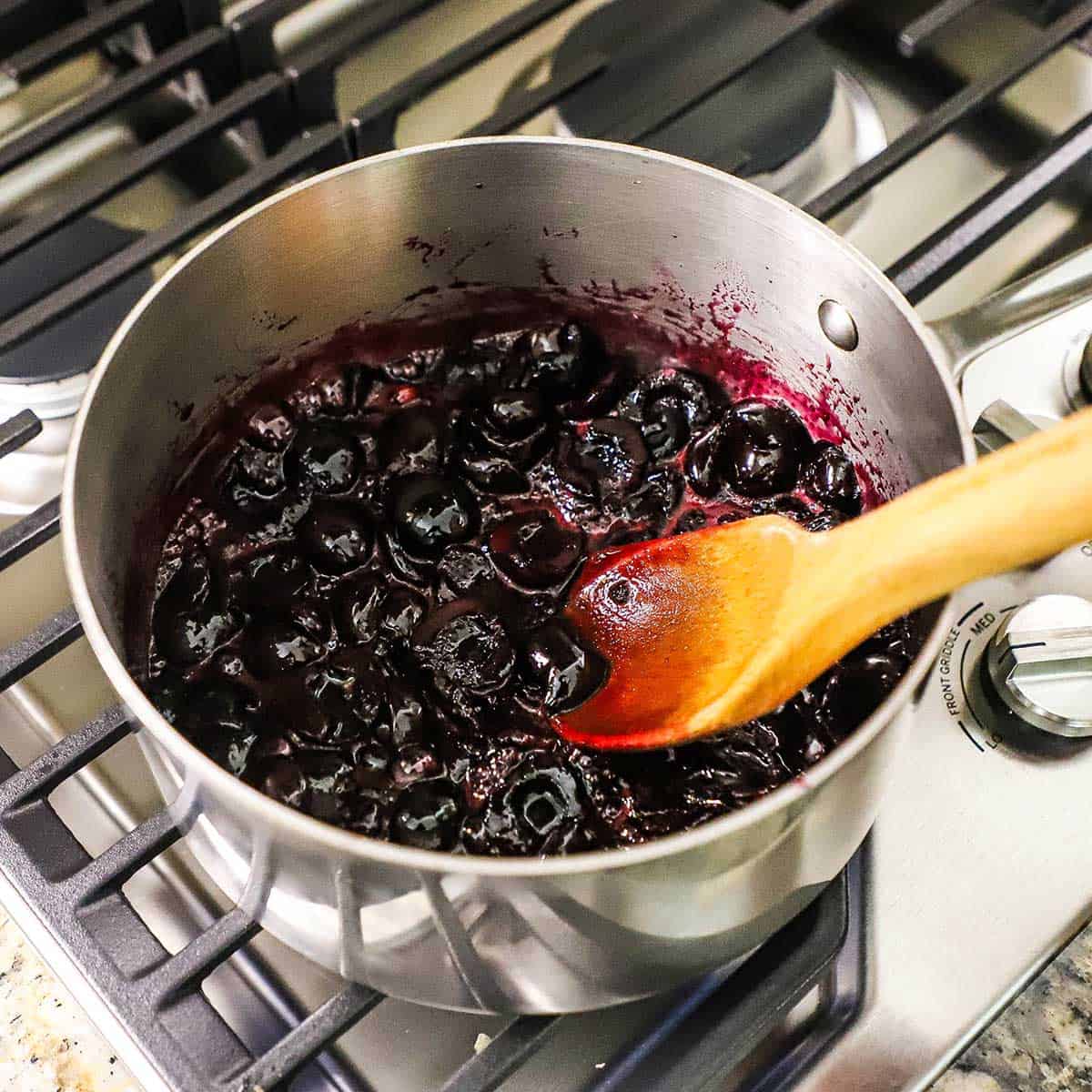 A person using a wooden spoon to stir a cherry fruit pie filling in a silver saucepan on a gas stove.