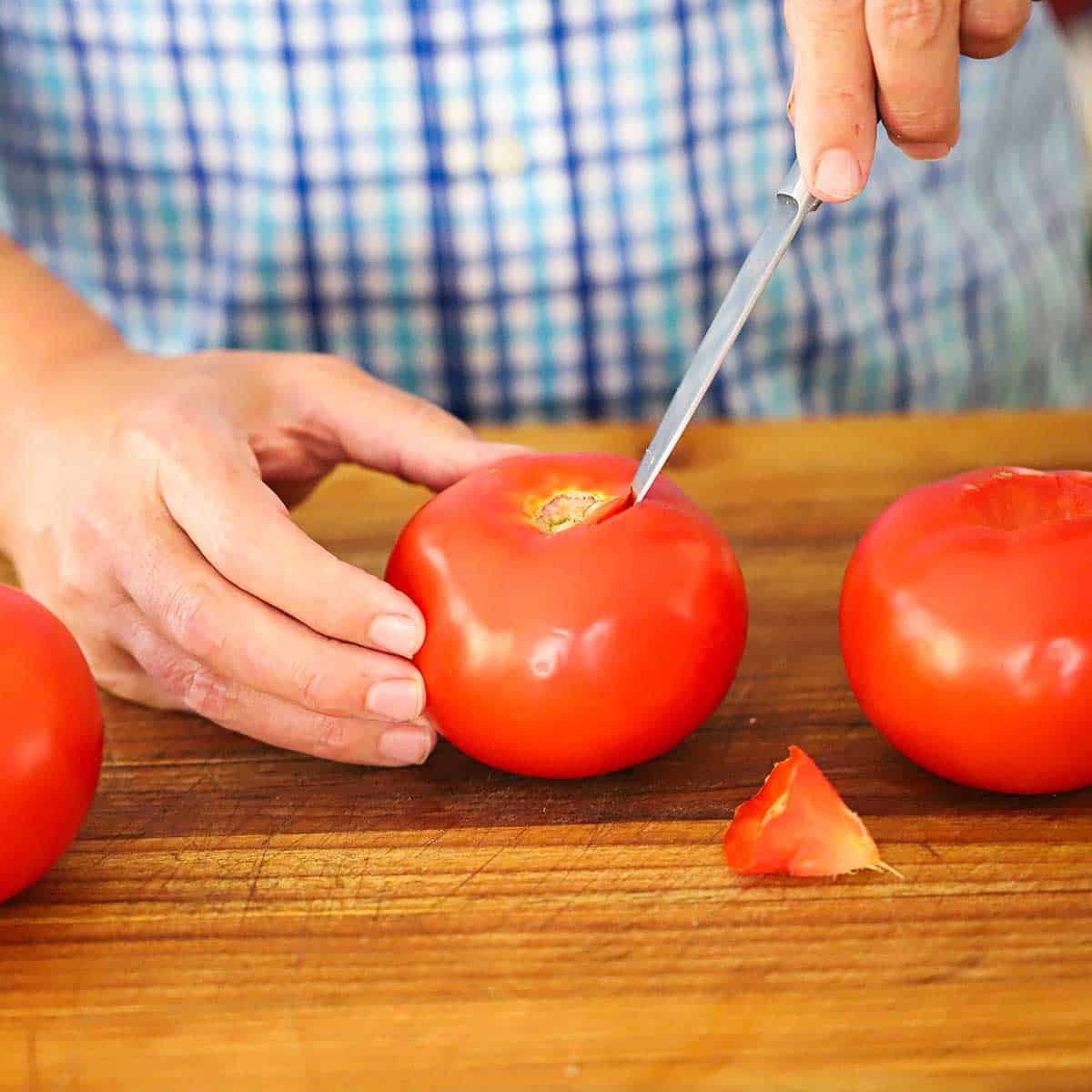 A person using a paring knife to cut the top stem portion out of a large red summer tomato on a wooden cutting board.