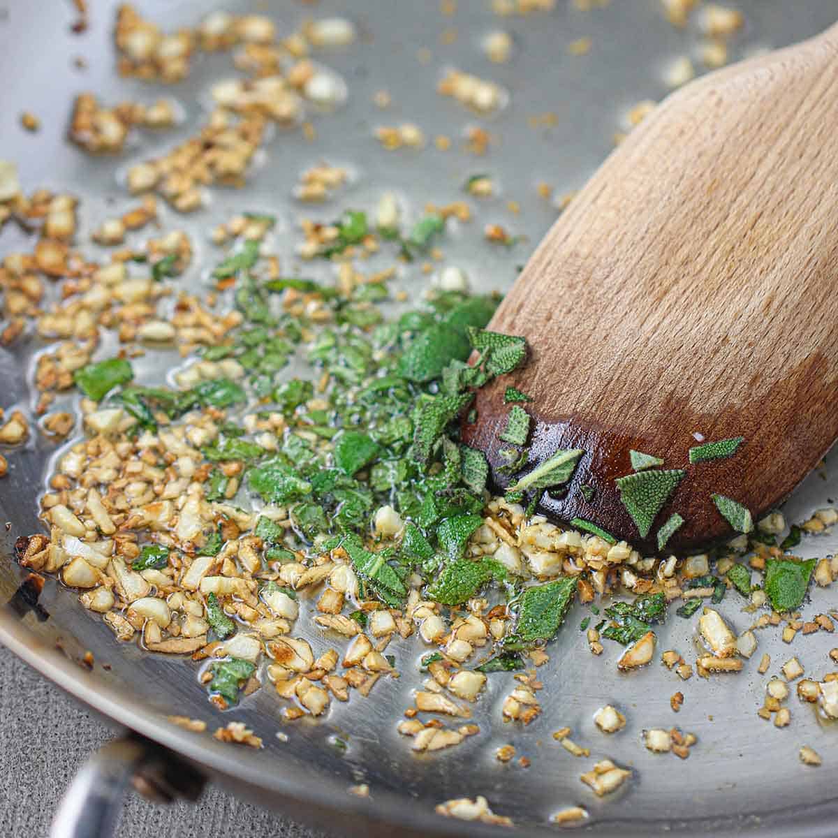 A person using a spoon to stir minced garlic and chopped fresh sage that is being sautéed in oil in a skillet.