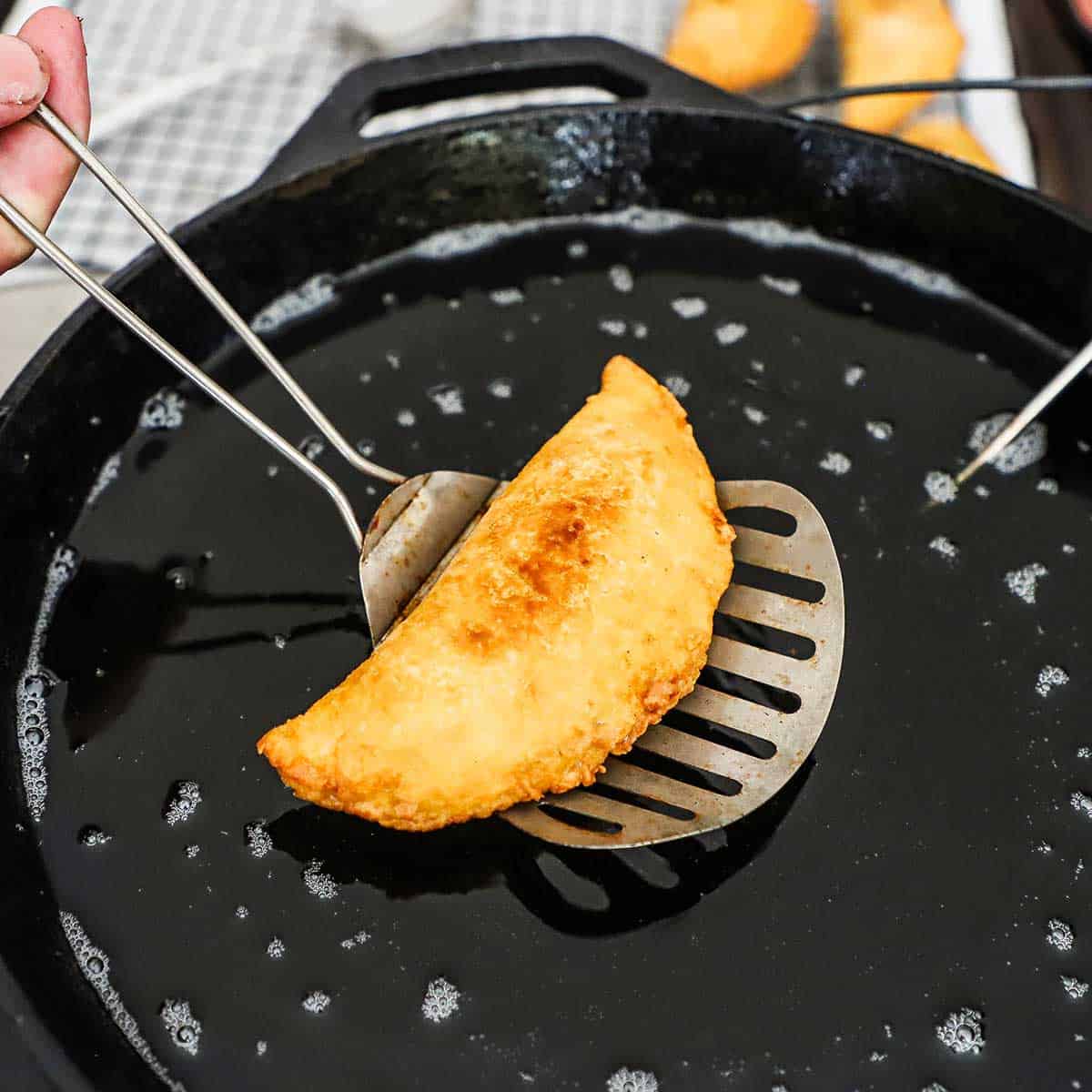 A person using a metal slotted spatula to raise a freshly Southern fried fruit pie from a skillet that is filled with hot oil.