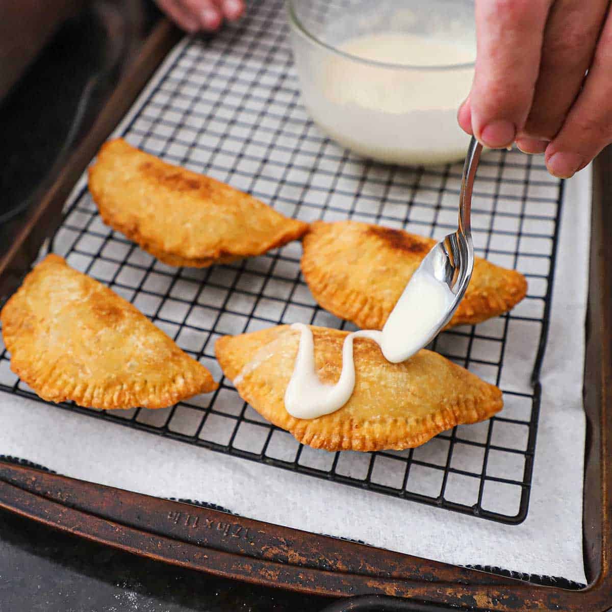 A person using a spoon to drizzle a sugar glaze over the top of a Southern fried fruit pie that is resting next to more pies on a baking rack.