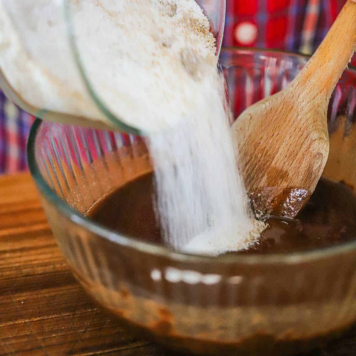 A person pouring flour and cocoa powder mixture into a glass bowl that is filled with melted butter combined with melted chocolate.