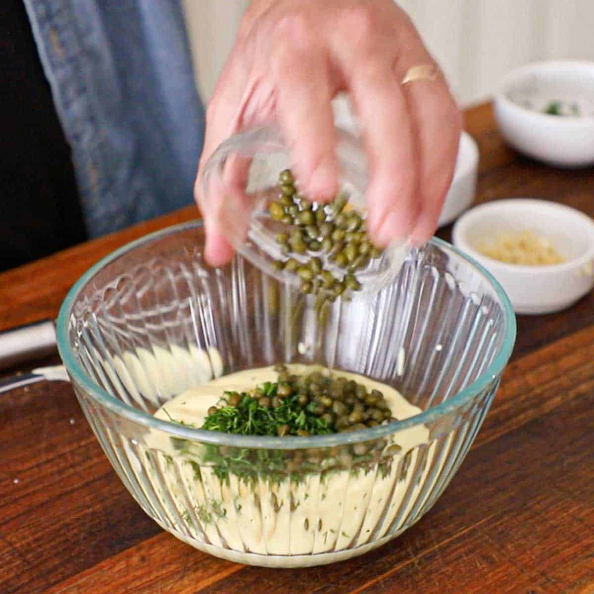 A person transferring capers from a small glass bowl into a large glass bowl filled with homemade mayonnaise, chopped dill, and Dijon mustard.