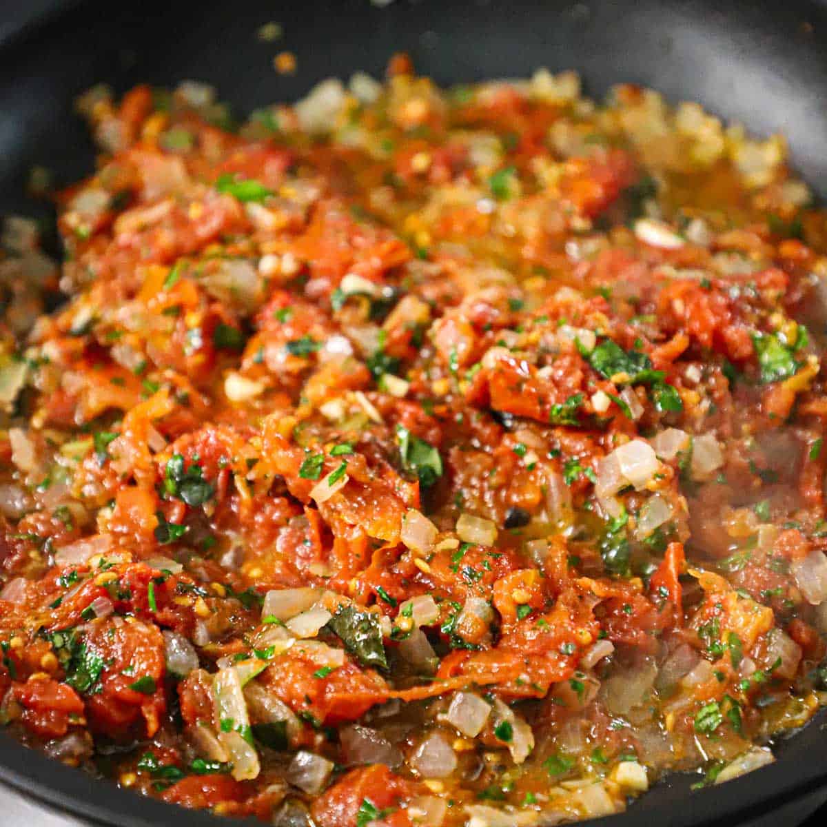 A close-up view of a crushed tomato, herbs, and onion sauce simmering in a large skillet on a gas stove.