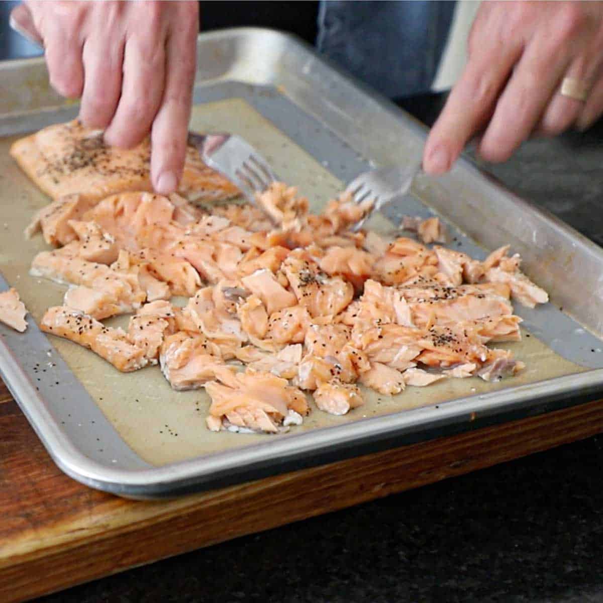 A person using two forks to tear baked seasoned salmon into chunks on a half sheet pan that has been lined with a silicon mat.