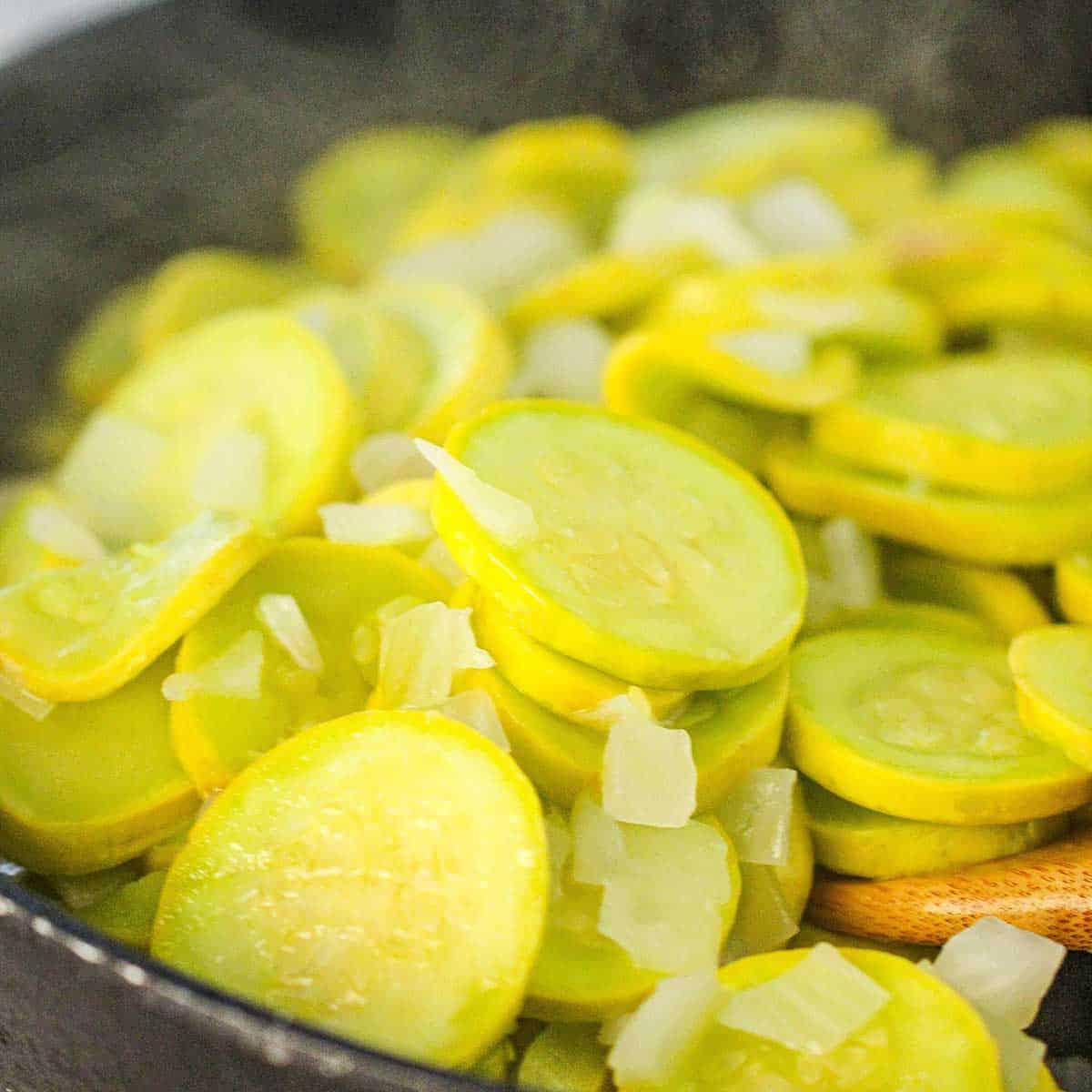 A close-up view of slices of yellow squash being sautéed with chopped onion in a large skillet.