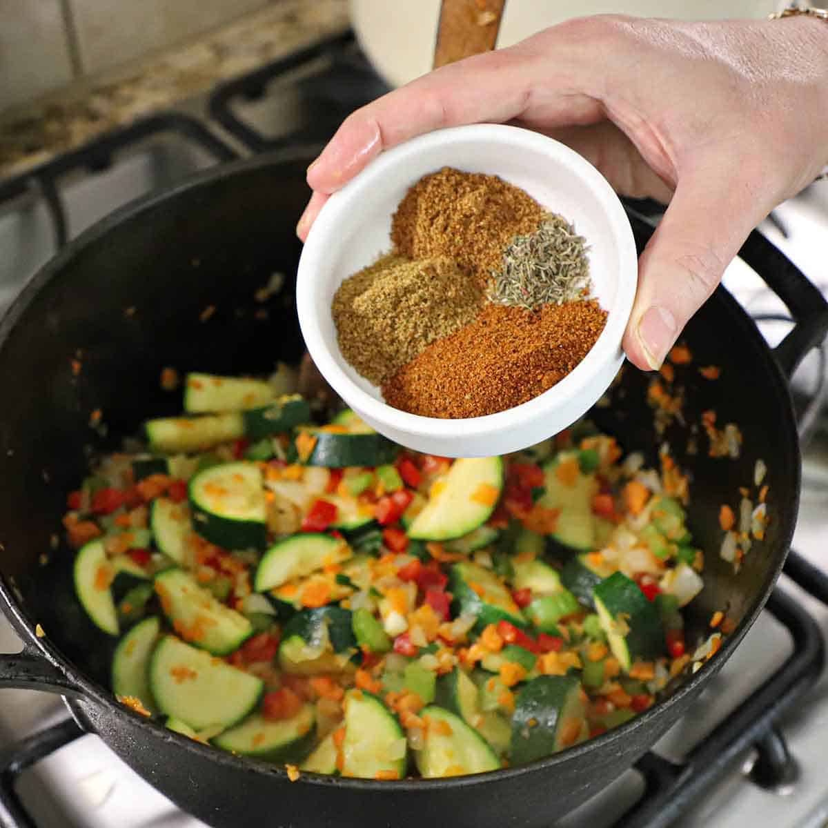 A person holding a small shallow white bowl containing small mounds of chili powder, ground cumin, dried oregano, and cayenne pepper over a cast-iron skillet filled with sautéed vegetables.