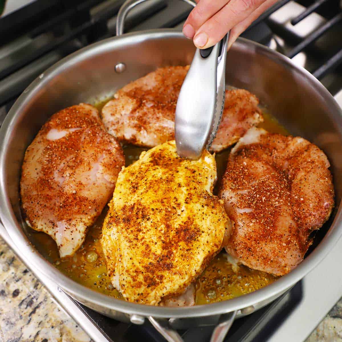 A person using a pair of metal tongs to flip a seasoned chicken breast that is being seared in a large skillet along with three more breasts on a gas stove.