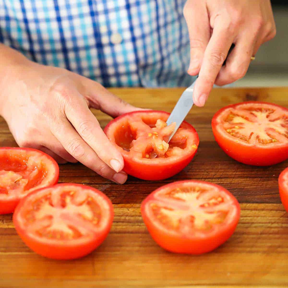 A person using a paring knife to cut the seeds from the middle of red tomatoes that have been cut in half and are resting cut-side up on a cutting board.