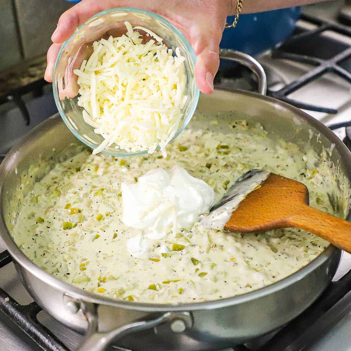 A person pouring shredded Monterey Jack cheese from a glass bowl into a skillet that is filled with a creamy white enchilada sauce.
