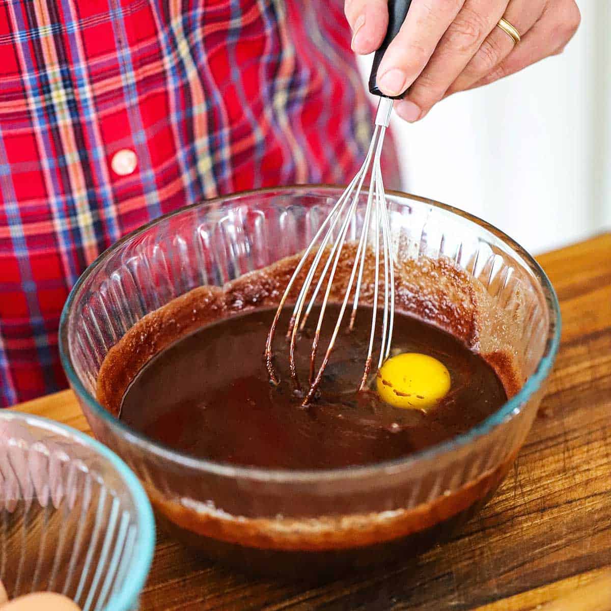 A person whisking a fudge brownie batter with a whole egg in a glass bowl on a wooden cutting board.