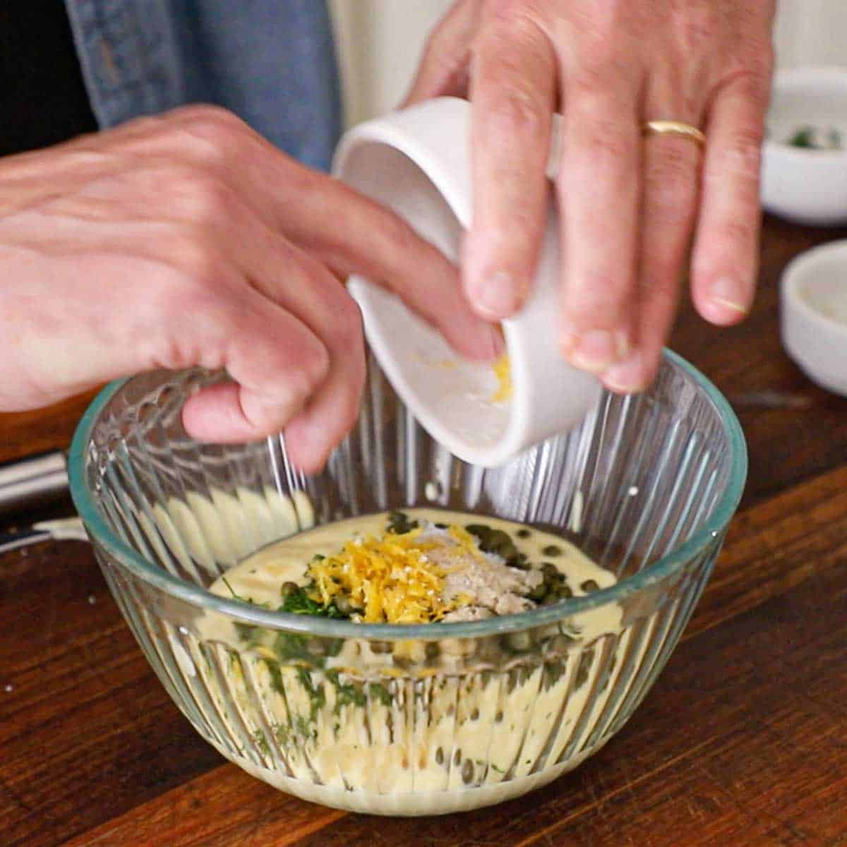 A person using his fingers to transfer lemon zest from a small white bowl into a glass bowl filled with mayonnaise, garlic, dill, capers, and lemon juice.