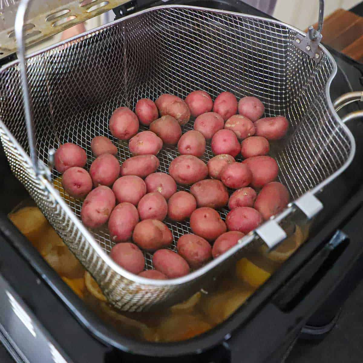 A person lowering a large basket filled with baby red potatoes into a turkey deep-fryer that is filled with boiling spiced water.