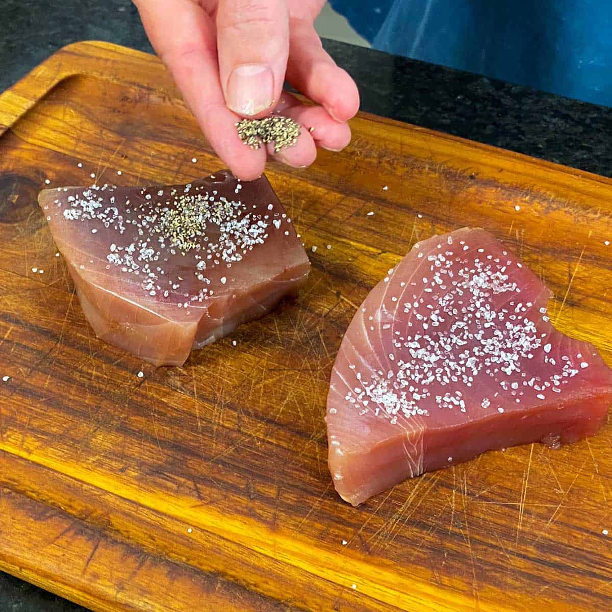 A person using his fingers to sprinkle salt and pepper on two raw tuna steaks that are resting on a wooden cutting board.