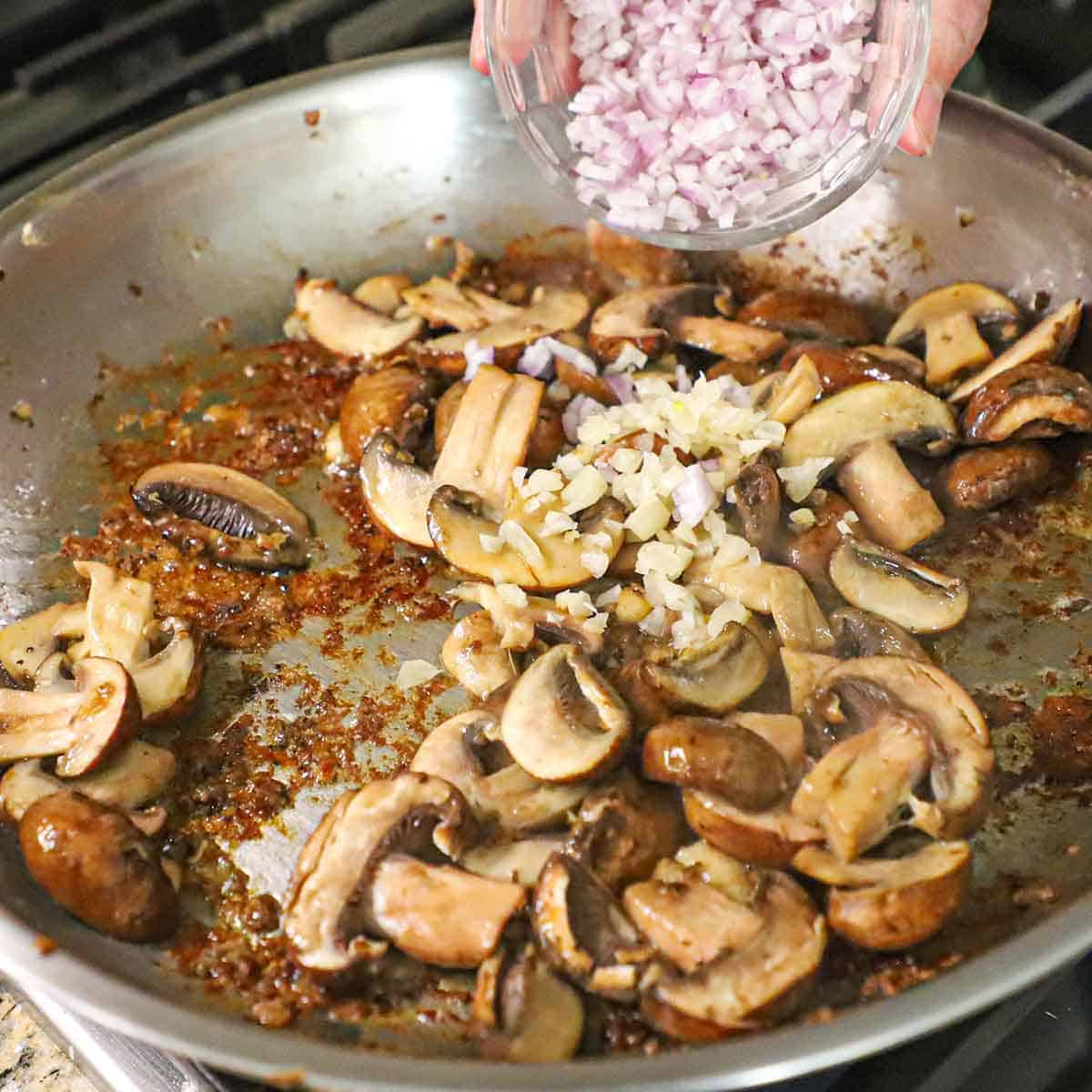 A person adding chopped shallots into a skillet filled with sautéed sliced baby Bella mushrooms and garlic.
