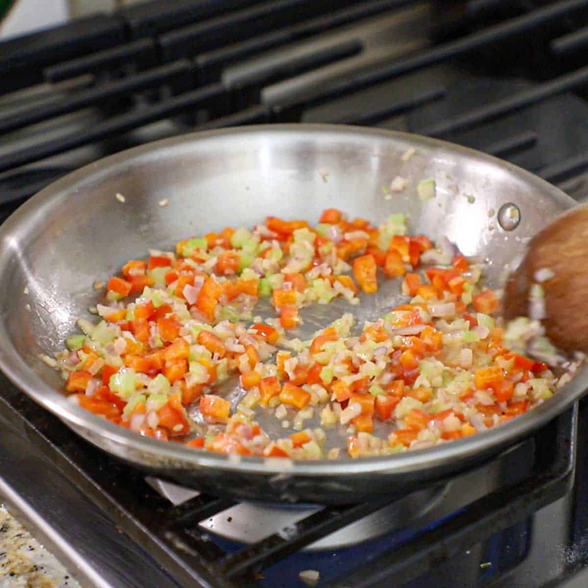 A person sautéing chopped red bell peppers, celery, and shallots in oil in a small saucepan on a gas stove.