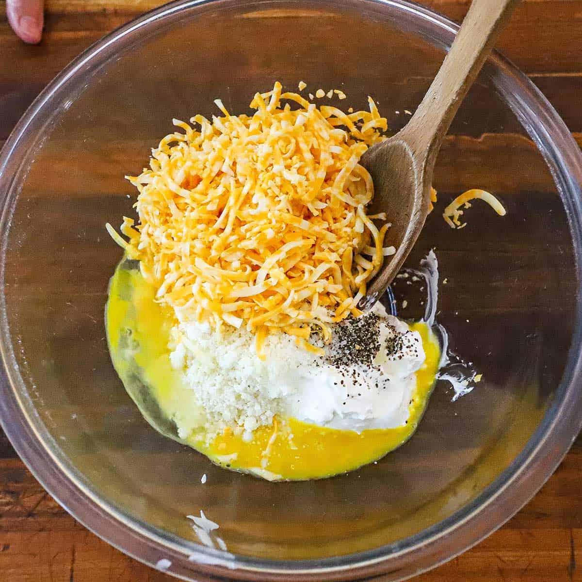 An overhead view of a large glass bowl filled with sour cream, mayonnaise, cheddar cheese, an egg, Parmesan cheese, and seasonings.