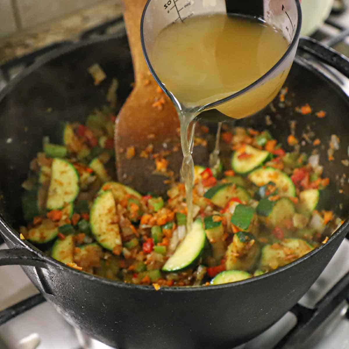 A person pouring vegetable stock from a measuring cup into a large cast-iron pot filled with seasoned sautéed vegetables.