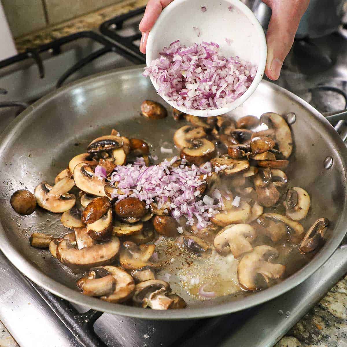 A person transferring chopped shallots from a small bowl into a large skillet that is filled with sautéed baby bella sliced mushrooms on a gas stove.