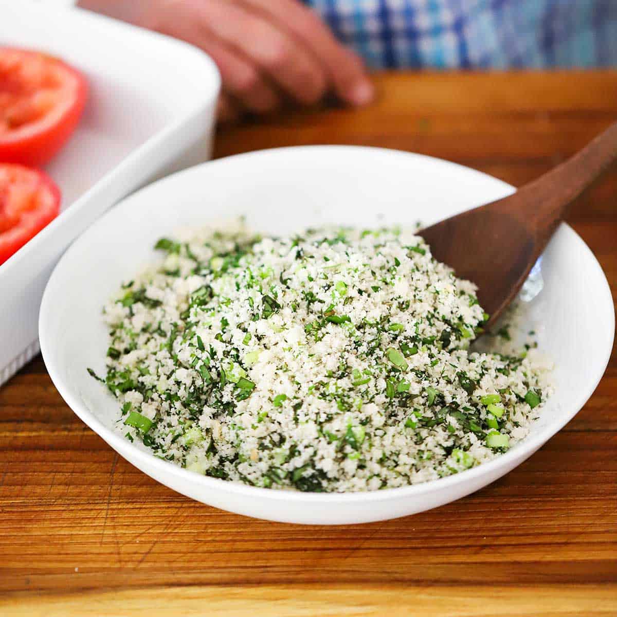 A person using a wooden spoon to mix breadcrumbs, chopped scallions, fresh herbs, garlic, and salt in a shallow white bowl.