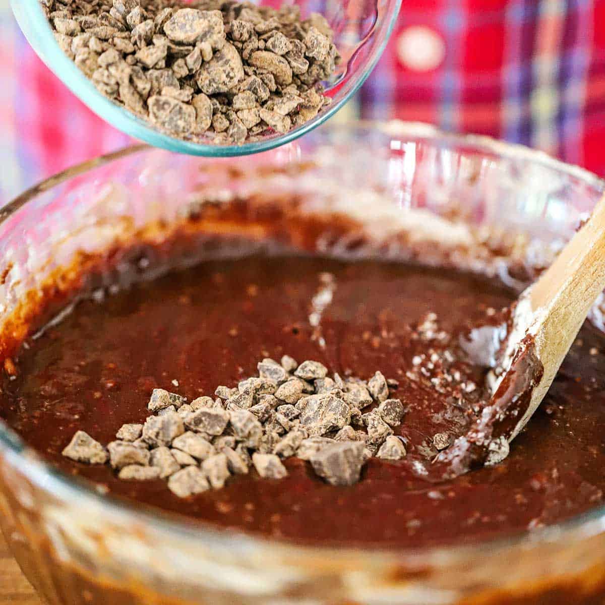 A person pouring chunks of semi-sweet chocolate into a large glass bowl that is filled with a fudge brownie batter.