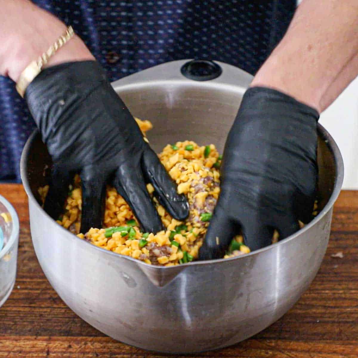 A person wearing black kitchen gloves working cubed high-temperature cheddar cheese and finely chopped jalapeño into a ground meat mixture for homemade Texas sausage.