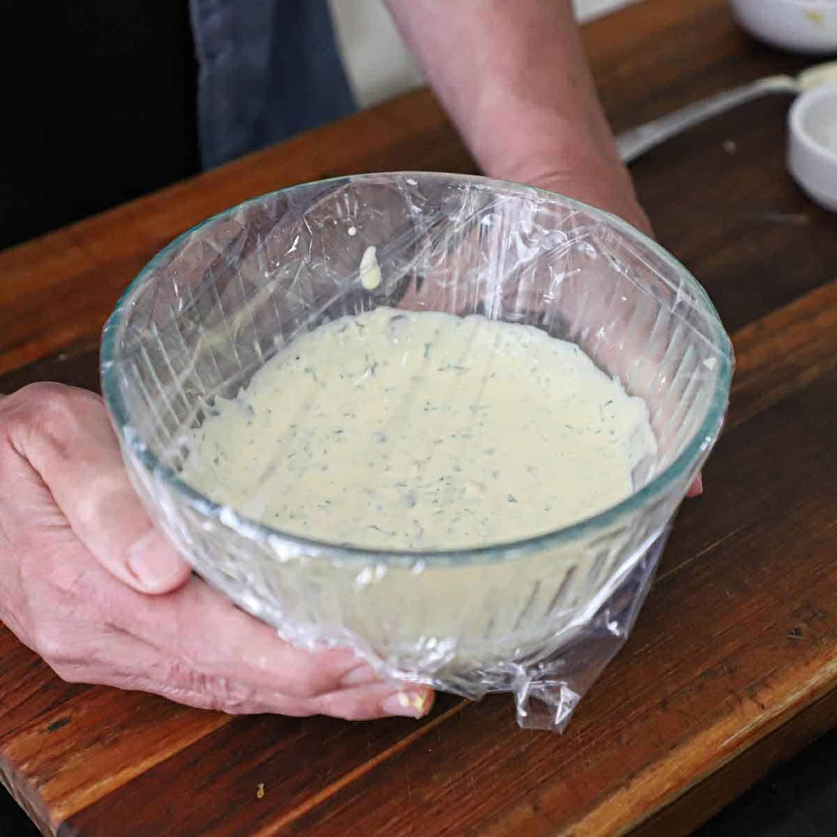 A person holding a glass bowl that is filled with freshly prepared Lemon-Dill and Caper Aioli with plastic wrap over the top of the bowl.