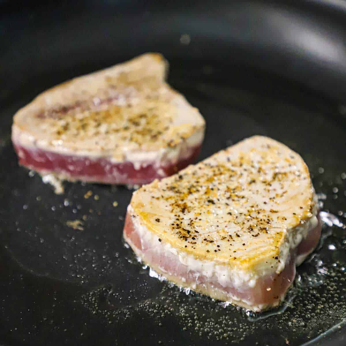 Two partially cooked seasoned tuna steaks being seared in a large black non-stick skillet in shimmering oil.