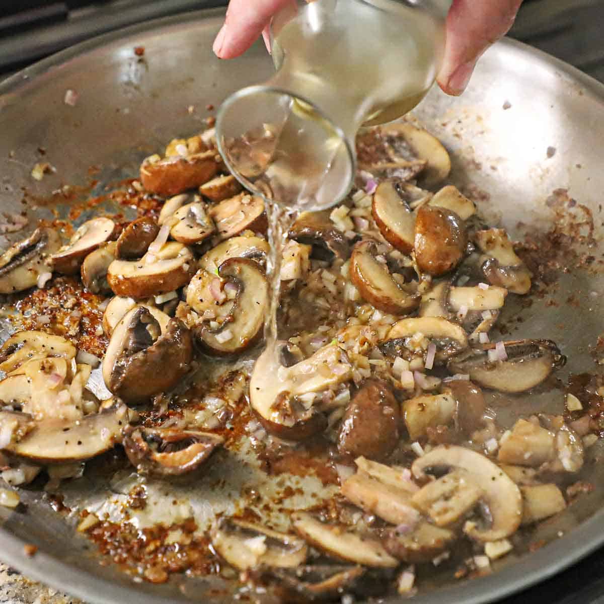 A person pouring white wine from a small carafe into a skillet filled with sautéed sliced mushrooms, shallots, and garlic.