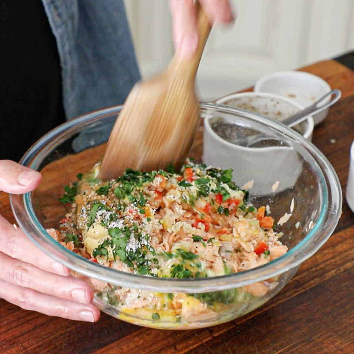 A person using a large wooden spatula to mix all the components for fresh salmon cakes in a large glass bowl on a wooden cutting board.